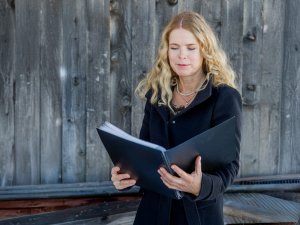Frau liest in einem Ordner in einem alten Gebäude Eine Frau steht vor einer Holzwand. Sie hält einen Ordner und schaut hinein. Es ist Tag und das Licht ist hell.