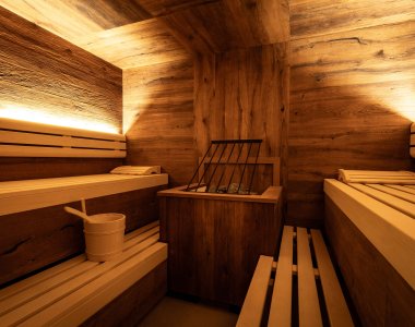 In einem Raum stehen Holzbänke mit einem Ofen. Eine Wasserschaufel und ein Eimer liegen bereit für die Sauna.