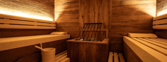 In einem Raum stehen Holzbänke mit einem Ofen. Eine Wasserschaufel und ein Eimer liegen bereit für die Sauna.