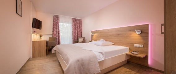 Das Zimmer hat ein großes Bett, einen Schreibtisch und einen Fernseher. Tageslicht kommt durch die Fenster.