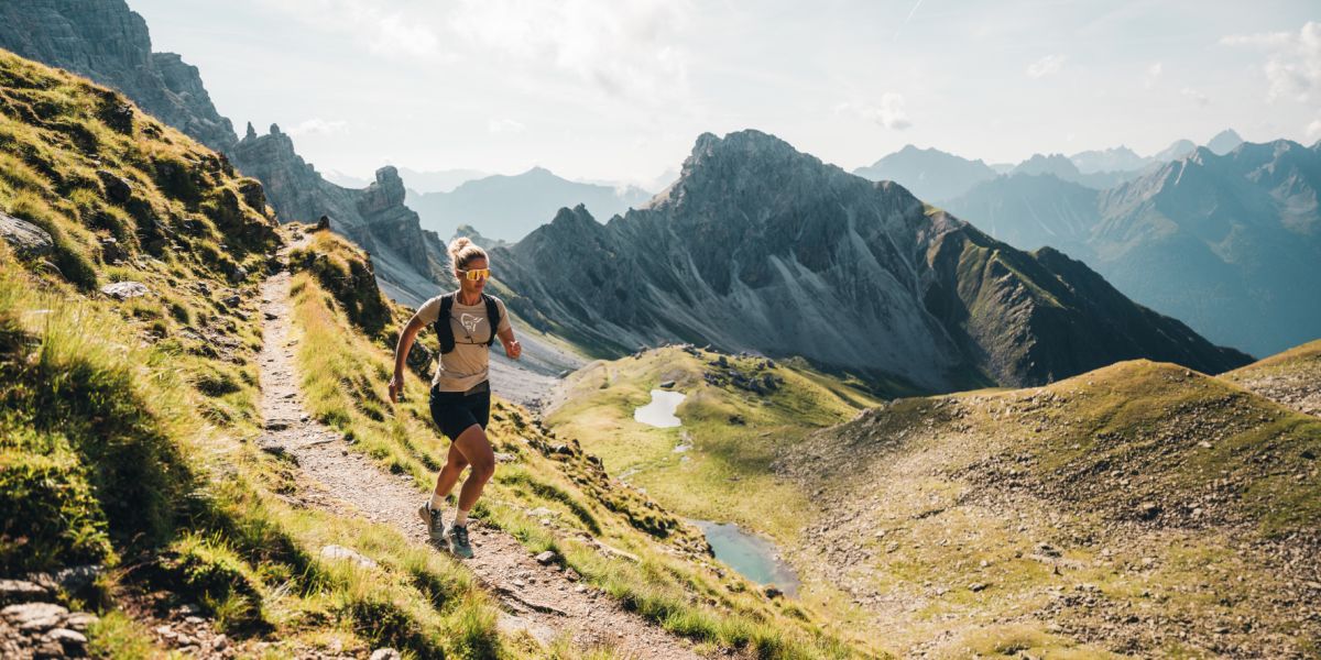 Eine Trailrunnerin rennt einen schmalen Bergpfad entlang, im Hintergrund sind die Berge der Stubaier Alpen zu sehen.