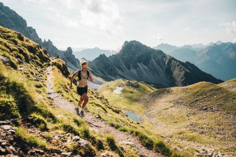 Un coureur de trail s'élance sur un sentier de montagne étroit ; à l'arrière-plan, on aperçoit les montagnes des Alpes de Stubai.