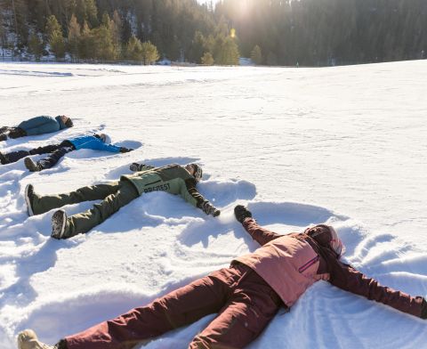 Familie liegt im Schnee und macht Schneeengel auf einer sonnigen Winterwiese im Ötztal