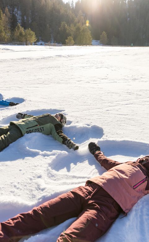 Une famille est allongée dans la neige et fait des anges dans une prairie hivernale ensoleillée de la vallée d'Ötztal.