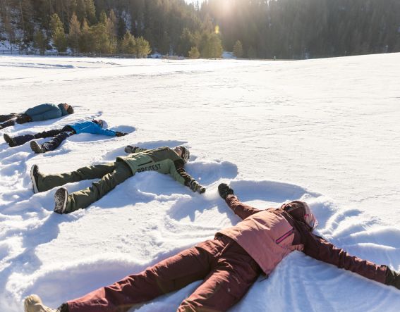 Familie liegt im Schnee und macht Schneeengel auf einer sonnigen Winterwiese im Ötztal
