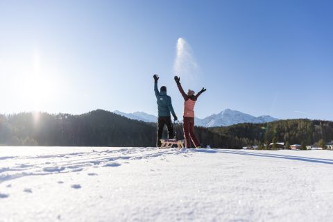 Paar jubelt neben einem Holzschlitten auf einer sonnigen Schneefläche im Ötztal – Wintermoment mit Bergblick