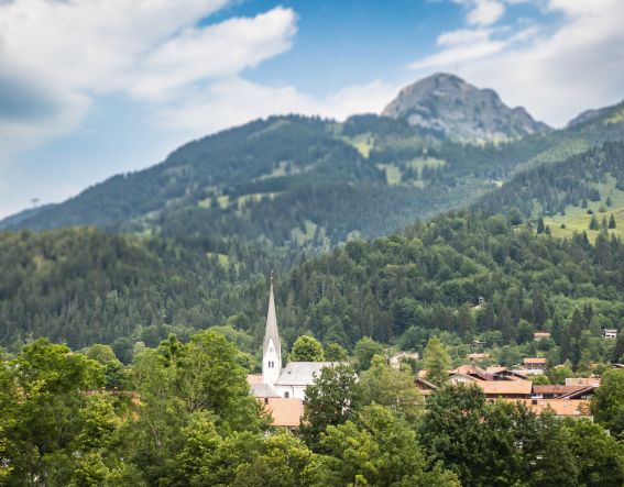 Bayrischzell liegt zwischen den Bergen und zwischen Bäumen schaut die Kirche hervor.