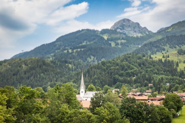 Bayrischzell liegt zwischen den Bergen und zwischen Bäumen schaut die Kirche hervor.