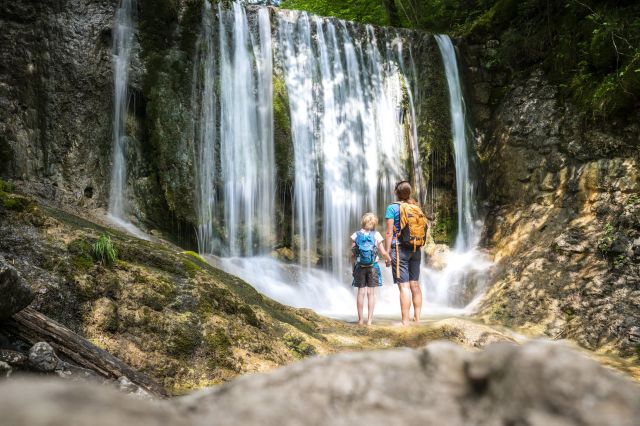 Mutter und Tochter schauen sich einen Wasserfall an. Ein Pärchen schaut sich einen Wasserfall an.