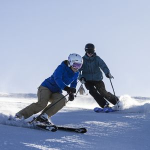 Zwei Personen fahren bei Sonnenschein den Sudelfeld mit den Ski hinunter.
