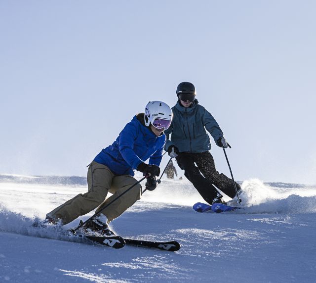 Zwei Personen fahren bei Sonnenschein den Sudelfeld mit den Ski hinunter.