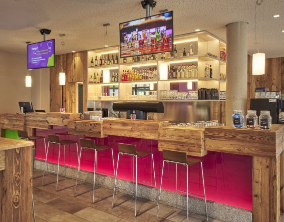 Moderne Hotelbar mit auffälligen roten Barhockern, Holzoberflächen und gut sichtbarem Getränkeangebot.