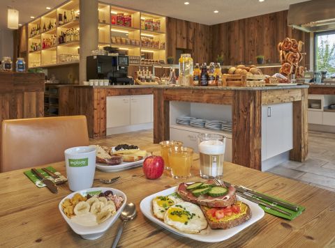 Un tavolo per la colazione apparecchiato di fronte al buffet con muesli, frutta fresca, caffè e succhi di frutta.
