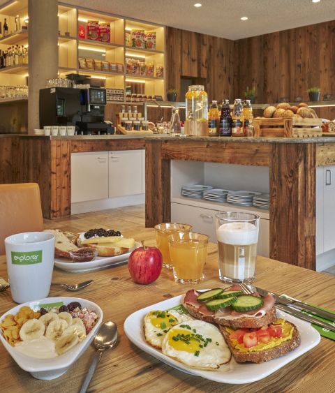 Gedeckter Frühstückstisch vor dem Buffet mit Müsli, frischem Obst, Kaffee und Säften.
