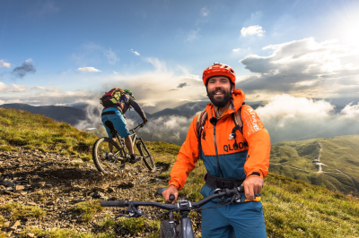 Zwei Personen fahren mit dem Mountainbike in den Bergen. Es ist sonnig und es gibt Wolken am Himmel.