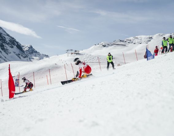 Ein Skiwettbewerb findet in den Bergen statt, viele Fahrer sind auf der Piste. Zuschauende stehen an der Strecke und beobachten.