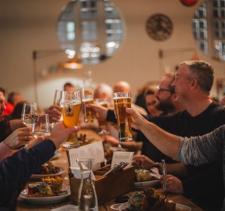 Eine Gruppe Menschen stößt in geseliger Runde am Tisch in der Fasshale mit ihren Getränken an