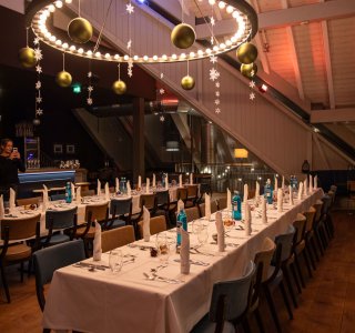 Eine lange Tafel ist für eine Veranstaltung im Advent gedeckt. Stühle sind aufgereiht, und dekorative Kugeln und Schneefocken hängen in Gold von der Lampe.
