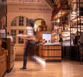 Momntaufnahme einer Servicemitarbeiterin, die Essen rausträgt. Im Hintergrund ist dei Bar zu sehen, an der ein Barkeeper arbeitet