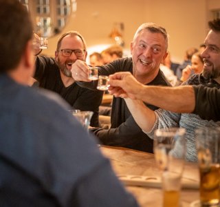 Vier Männer stoßen mit Schnapsgläsern an und lachen. Auf dem Tisch steht Bier und außenrum hängt Weihnachtsdekoration