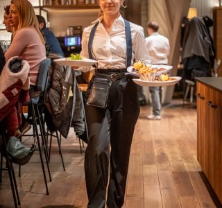 Eine Kellnerin geht durch die Fasshalle und bringt Essen. Es ist Abend und viele Gäste sitzen.