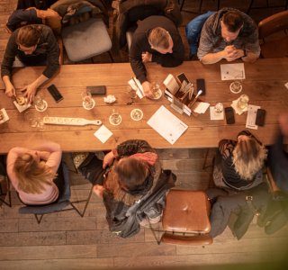 Viele Menschen sitzen an einem Tisch in der Fasshalle und genießen die Zeit zusammen. Sie trinken und unterhalten sich.