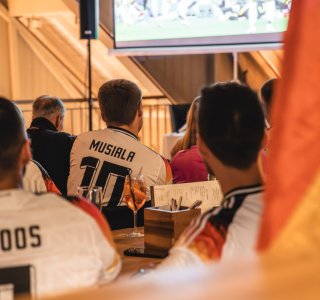 Die Menschen schauen ein Fußballspiel in der Fasshalle an und tragen Trikots. Stimmung ist lebhaft und konzentriert.