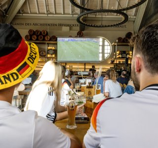 Fans schauen das Fußballspiel von Deutschland in der Fasshalle. Getränke stehen auf dem Tisch. Die Stimmung ist lebhaft und aufregend.