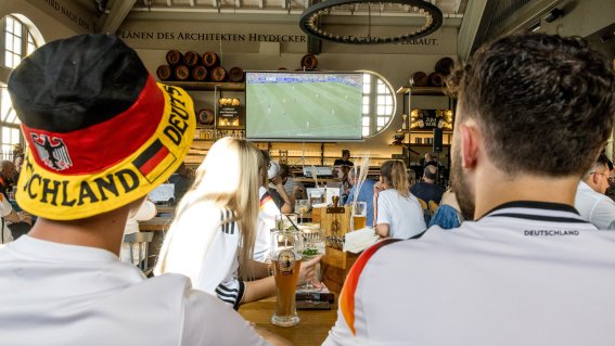 Fans schauen das Fußballspiel von Deutschland in der Fasshalle. Getränke stehen auf dem Tisch. Die Stimmung ist lebhaft und aufregend.