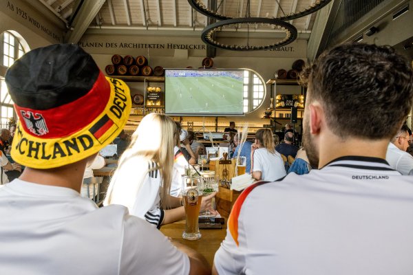 Fans schauen das Fußballspiel von Deutschland in der Fasshalle. Getränke stehen auf dem Tisch. Die Stimmung ist lebhaft und aufregend.