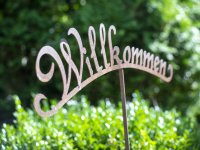 Ein Willkommensschild steht in einem Garten zwischen grünen Büschen. Es zeigt den Text Willkommen in geschwungener Schrift.