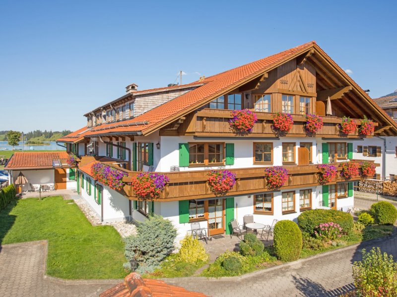 Das Gebäude steht in Bayern. Es hat Balkone mit Blumen und eine grüne Landschaft. Tageslicht scheint auf die Fassade.