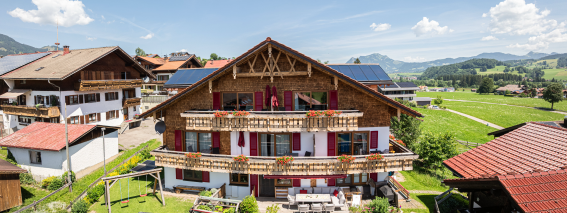Das Haus steht in den Alpen. Es ist Sommer und die Sonne scheint. Menschen sitzen draußen und genießen die Aussicht.