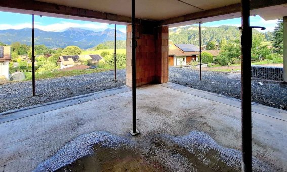 Der Innenraum eines Rohbaus zeigt Stützen und einen weiten Blick auf eine grüne Landschaft und Berge im Hintergrund.