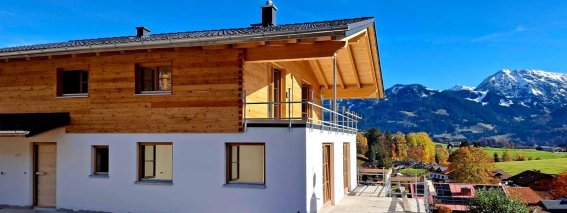 Ein neues, helles Haus steht in der Nähe von Bergen unter blauem Himmel. Die Umgebung ist grün und einladend.