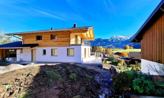 Ein neues Haus wird in einer ländlichen Gegend der Alpen gebaut. Vor dem Gebäude sind Pflanzen und Berge sichtbar.