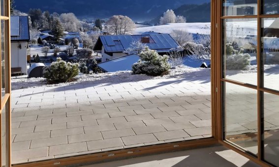 Durch ein offenes Fenster sieht man Schnee und Berge. Es ist tagsüber und die Sonne scheint.