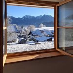 Blick auf einen verschneiten Bauernhof in den Bergen Durch ein offenes Fenster sieht man einen verschneiten Bauernhof und Berge bei klarem Himmel.