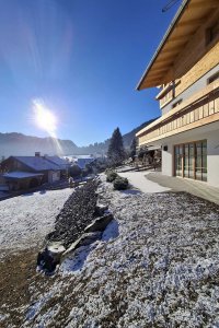 Der Schnee liegt auf dem Boden. Die Sonne scheint. Ein Haus steht in der Landschaft. Bäume sind im Hintergrund sichtbar.