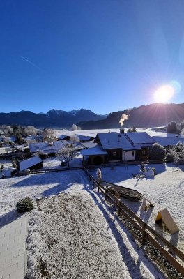 Ein Winterdorf zeigt sich am Morgen unter Schnee. Sonnenstrahlen scheinen auf das Dorf und die Berge im Hintergrund.