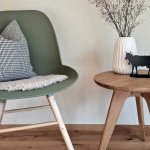 Stuhl mit Kissen und Tisch in einem Raum Ein Stuhl steht neben einem Tisch mit einer Blumenvase und einer Figur. Der Raum hat einen hellen Boden und Wände.