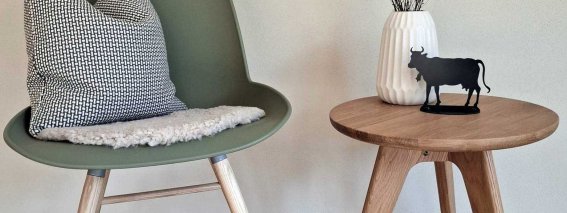 Ein Stuhl steht neben einem Tisch mit einer Blumenvase und einer Figur. Der Raum hat einen hellen Boden und Wände.