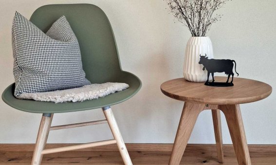 Ein Stuhl steht neben einem Tisch mit einer Blumenvase und einer Figur. Der Raum hat einen hellen Boden und Wände.