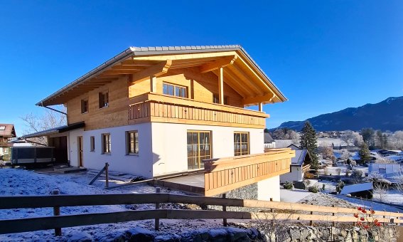 Ein Holzhaus steht in einer verschneiten Landschaft. Der Himmel ist blau. Es gibt Berge im Hintergrund.