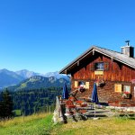 Die Hütte steht in den Bergen. Menschen sitzen draußen und genießen die Aussicht auf die Landschaft und den blauen Himmel.