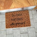 Die Matte liegt vor einer Tür und begrüßt Besucher mit einem Text. Es sind schwarze Punkte auf dem braunen Material.
