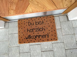 Die Matte liegt vor einer Tür und begrüßt Besucher mit einem Text. Es sind schwarze Punkte auf dem braunen Material.