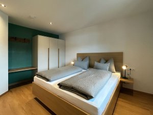 Das Zimmer zeigt ein Doppelbett mit zwei Kissen und einer hellen Wand. Es gibt einen Kleiderschrank und eine Ablage.