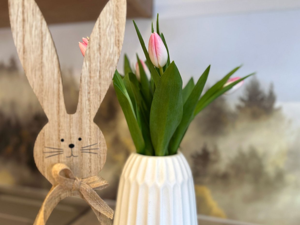 Ein Holzhase steht neben einer Vase mit Tulpen auf einem Tisch. Der Frühling ist da. Die Sonne scheint durch das Fenster.