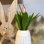 Holzhase und Frühlingsblumen auf einem Tisch Ein Holzhase steht neben einer Vase mit Tulpen auf einem Tisch. Der Frühling ist da. Die Sonne scheint durch das Fenster.